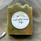 A rectangular bar of handmade soap in a muted olive green color, wrapped in a sheer white organza bag with a white ribbon bow, and featuring a round white label. The soap is laid flat on a textured brown kraft paper background. Natural Basil & Lemongrass Soap. 