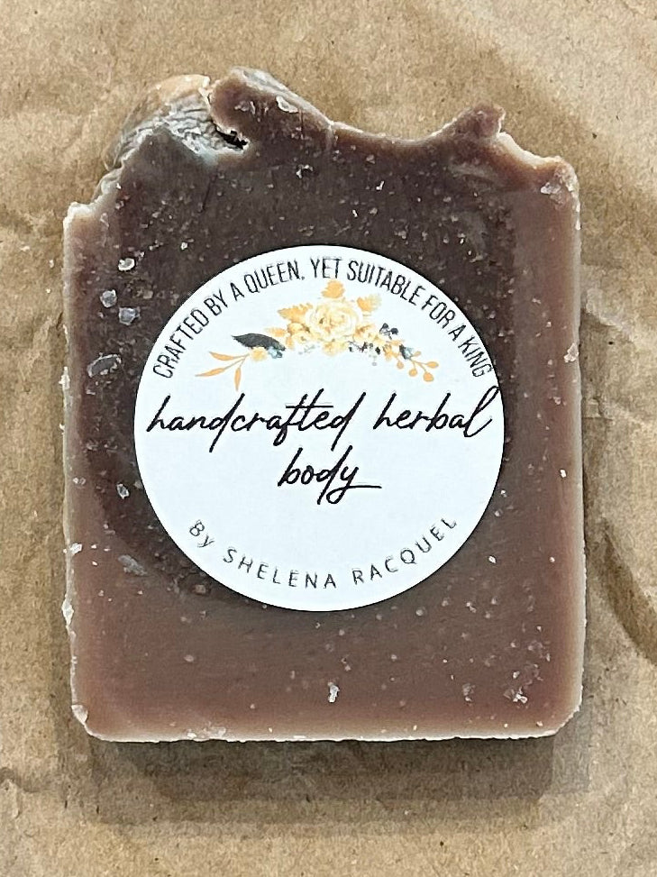 A small rectangular bar of soap with a clear top and a round white label reading 'handcrafted handmade soap' by The Soap Company, placed on top of crumpled brown paper over a tan surface.
