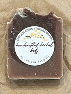 A small rectangular bar of soap with a clear top and a round white label reading 'handcrafted handmade soap' by The Soap Company, placed on top of crumpled brown paper over a tan surface.