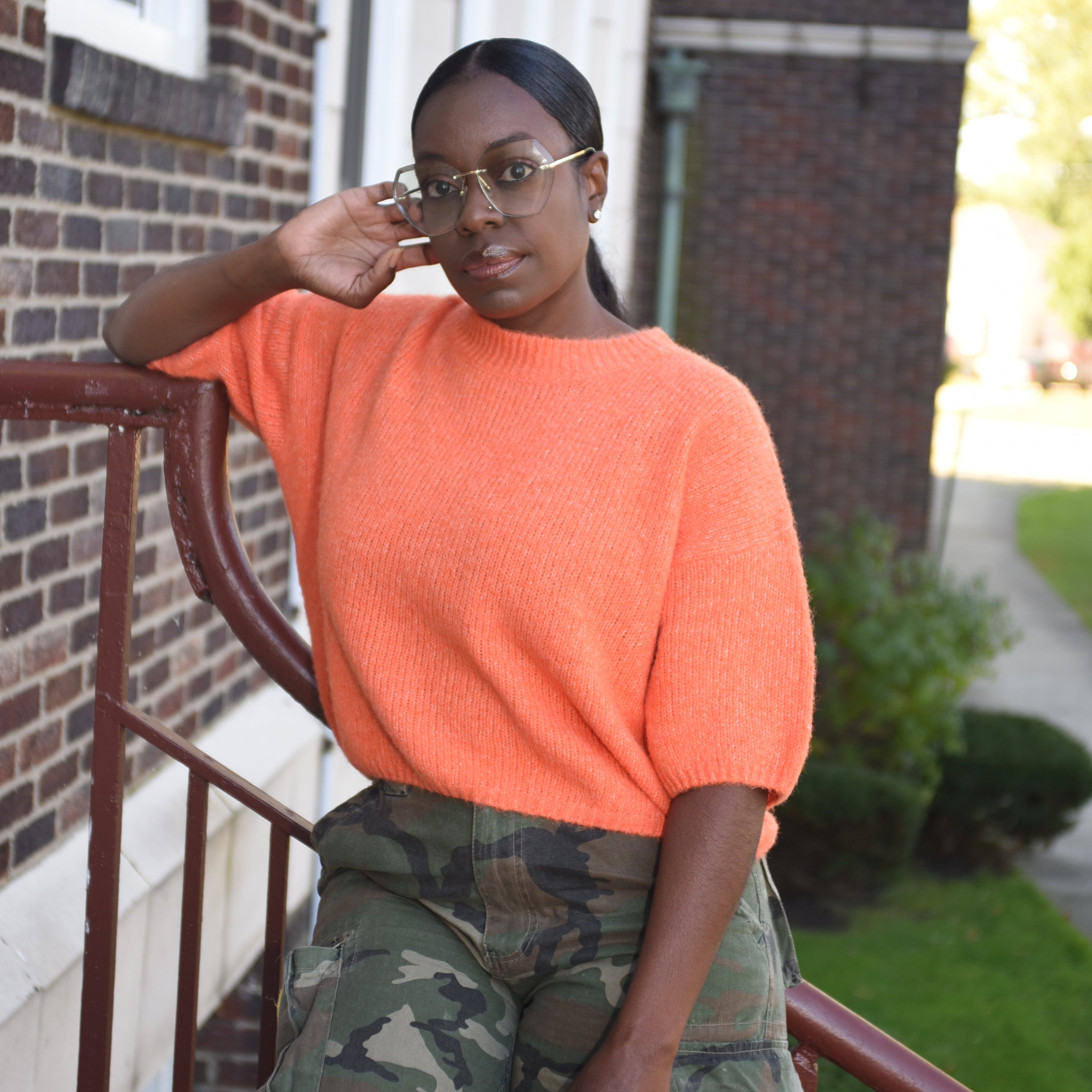 Person wearing an orange shirt and camouflage pants sitting on a chair outdoors.